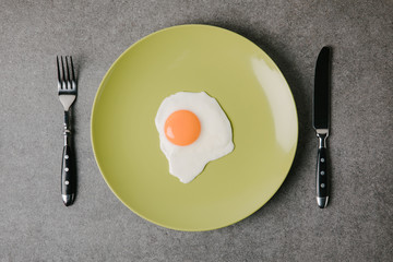 top view of tasty fried egg on plate and fork with knife on grey