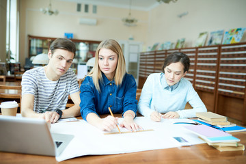 Fototapeta premium Serious confident student friends examining blueprint in library: concentrated guy pointing at laptop screen and explaining rules to groupmate