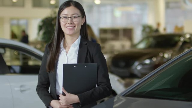 PAN of Asian saleswoman with clipboard smiling and posing for camera at car dealership