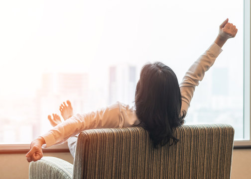 Life quality concept with business woman rear view relaxing sitting in rest on cozy armchair in modern hotel guest room or luxury home living room looking toward beautiful city urban scene