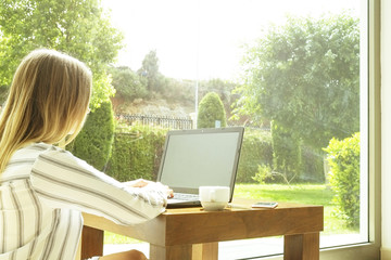 Young fit attractive woman sitting on floor, coffee table near big garden view window, working, browsing, surfing, blogging on laptop computer, blank screen. Freelance concept. Background, copy space