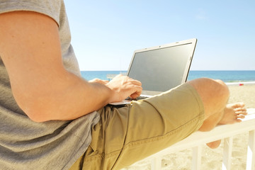 Fit young man using laptop sitting on white wood beach veranda porch. Perks of freelance concept. Male programmer working coding on notebook computer, blank screen. Sunny ocean sea. Copy space backgro
