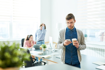 Serious thoughtful handsome young office employee drinking fresh coffee and reading sms on smartphone while his colleagues finding solution in background