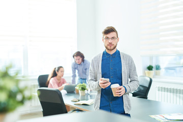 Portrait of calm confident successful young male designer with beard using gadget and drinking coffee in modern office