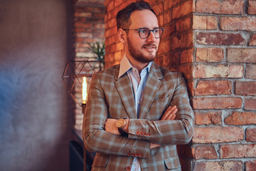 Fototapeta na wymiar Stylish man in a flannel suit and glasses with crossed arms leaning against a brick wall in a room with loft interior.