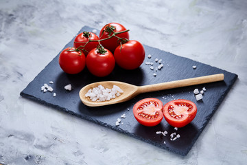 Fresh tomatoes and a spoon of salt on black stony board over white background, close-up, selective focus.