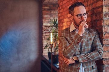 Portrait of a stylish man in a flannel suit and glasses leaning against a brick wall in a room with loft interior.