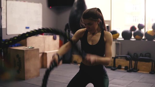 Strong sportive brunette girl in her 20's performing battle ropes workout at the gym.