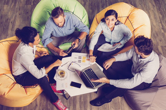 Four Content Young Business People Sitting On Beanbag Chairs Around Small CafÃ© Table, Working And Discussing Issues. Woman Is Looking At Camera And Smiling. High Angle View.