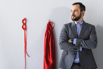 super businessman standing with crossed arms near mask and cape on wall in office