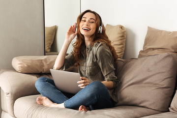 Obraz premium Image of pleased woman 20s with brown hair listening to music using wireless headphones and laptop, while sitting on sofa in living room with legs crossed