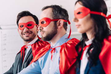 side view of smiling super businesspeople in masks and capes looking away in office