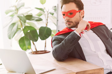 handsome super businessman in mask and cape looking at laptop in office