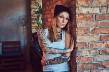 Naklejka premium A tattoed sexy blonde in a t-shirt and a hat posing against a brick wall.