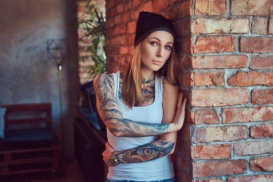 A tattoed sexy blonde in a t-shirt and a hat posing against a brick wall.