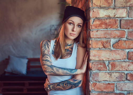 A tattoed sexy blonde in a t-shirt and a hat posing against a brick wall.