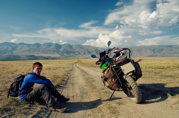 Motorcycle traveler man sitting on extreme rocky steppe road path in a mountain plateau in cloudy weather on the background of hills Altai Mountains Siberia Russia
