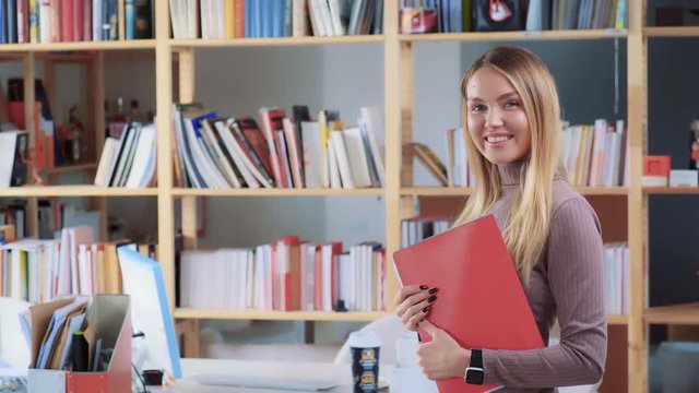 Beautiful long-haired blonde with a red folder for documents looking into the camera and smiling, showing white even teeth. An office worker with a beautiful smile looks at the camera, shot from above