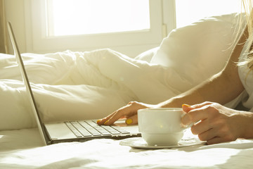 Close up, young blond woman in bed, white sheets linens holding cup of morning coffee, surfing browsing blogging eshopping on laptop. Perks of freelance concept. Blond female in tight shirt Background