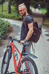 A handsome redhead male with a stylish haircut and beard dressed in sportswear and sunglasses walks in the park with a bicycle and backpack.