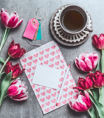 Cup of morning coffee with tulips flowers and blank paper card mock up with hearts, top view. Holidays breakfast. Flat lay style. Greeting card concept