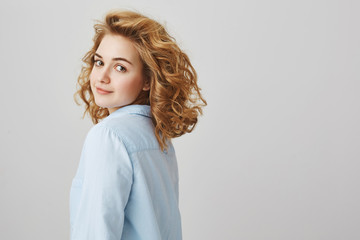 Studio portrait of good-looking slender woman with curly blonde hair turning and smiling at camera while stending in profile over gray background. Sensual girlfriend waits for her lover