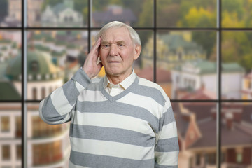 Portrait of senior man with head pain. Grandfather is suffering from headache, windows background with view on small european town.