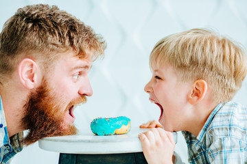 Side view portrait of beard red hair father and his son with open mouth share sweet donut between them.