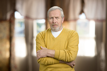 Fototapeta premium Sorrowful tragic old man portrait. Sad frustrated senior grandpa in yellow sweater with folded arms. Blurred room background.