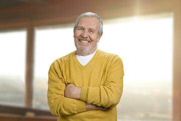 Portrait of smiling grandpa at home. Cheerful old man in yellow sweater with crossed folded arms against windows background indoor.