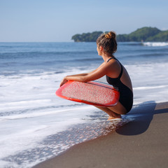 Naklejka na ściany i meble tanned surfer sitting with red surfboard on beach near ocean