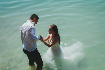 Wedding couple is holding hands in azure blue lake splashes of water. Beautiful bride in puffy...