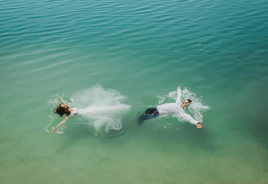 Wedding couple is swimming in azure blue lake splashes of water. Beautiful bride in puffy dress and groom having fun. Summer passion crazy emotions photo on seaside. Wet wedding clothes.