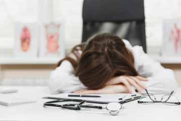 Young tired exhausted woman sitting at desk, working on computer with medical documents in light...