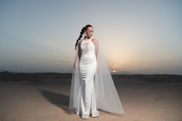 Fototapeta na wymiar Bride in wedding gown on sunset sky. Woman in white dress in desert. Sensual woman with brunette hair. Fashion model in sand dunes. Beauty girl with glamour look and makeup