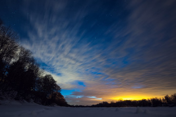 Snow-covered river near the forest on a winter frosty night with clouds and stars in the sky.