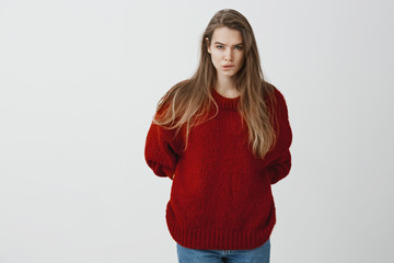 Studio shot of bothered concerned attractive european woman in red loose sweater, biting lip and frowning, staring at camera, holding hands behind, thinking, being worried and anxious over gray wall