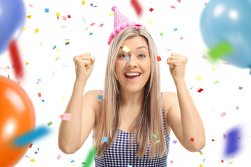 Young woman wearing a party hat gesturing happiness with confetti streamers and balloons flying around her