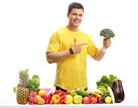 Young man behind a table with fruit and vegetables holding broccoli and pointing