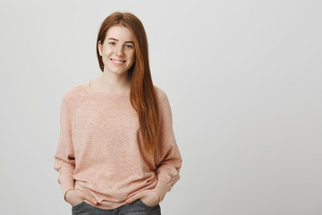 Portrait of adorable redhead caucasian woman standing with hands in pockets over gray background, smiling and expressing good-mood, wearing casual pullover. Peer asks guy to go to cinema