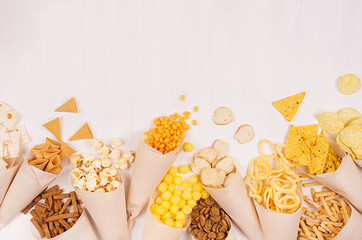 Beige paper cones with crunchy fast food snacks — nachos, popcorn, croutons, chips arranged on white wood board with copy space © finepoints