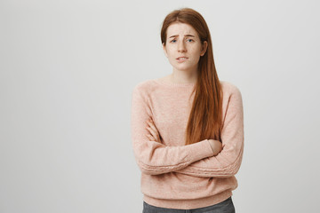 Gloomy unsatisfied caucasian redhead girl with freckles standing with crossed hands in defencive pose, feeling uncomfortable and anxious, over gray background. Woman worries for brother in hospital