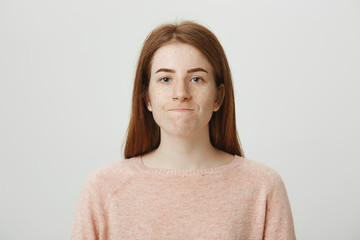 Close-up studio shot of annoyed or bothered redhead woman with stretched unhappy smile, standing over gray background. Girl is irritated by coworker and tries to hold her temper