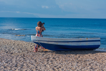 Obraz premium Beautiful female photographer in the dress with professional camera near boat on the sand