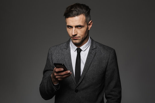 Image of serious man employer dressed in business costume using cell phone in office, isolated over gray background