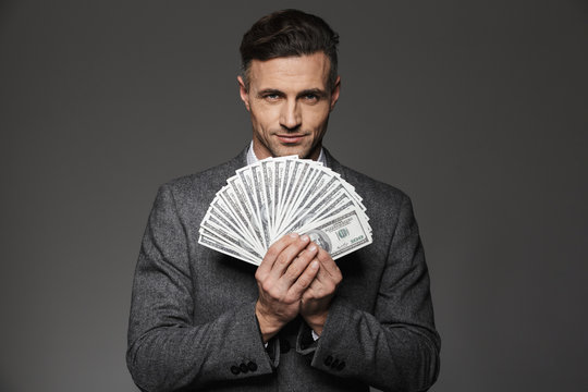 Photo of confident guy 30s in business suit holding fan of money dollar bills and looking on camera, isolated over gray background