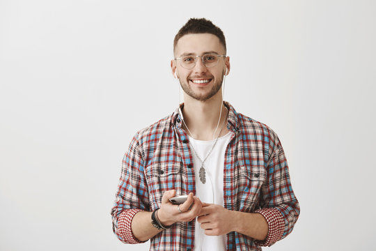 Favorite Music Helps Being Productive During Day. Portrait Of Handsome Happy Caucasian Guy In Glasses Holding Smartphone While Listening Music In Earphones, Smiling Broadly At Camera