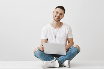 Boyfriend wants to show his couple something in laptop. Portrait of good-looking young bearded student in stylish clothes and glasses sitting with crossed legs on floor in campus holding notebook