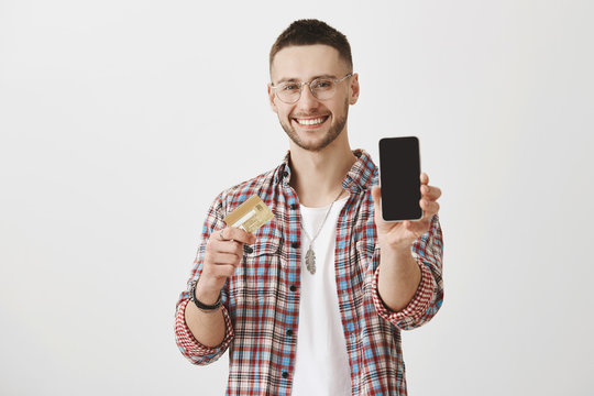 Grab your credit card and buy this phone. Portrait of good-looking pleased young man with bristle and eyewear, pulling hand towards camera and showing smartphone while holding bank card.