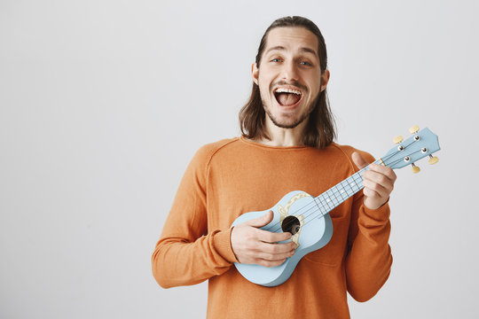 Showing His Feeling In Song. Portrait Of Happy Carefree Male Musician In Orange Sweater Playing Ukulele And Singing Out Loud, Being Satisfied And Pleased While Relaxing In Friends Circle